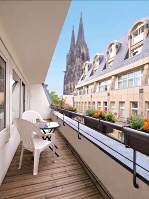 A balcony with a wooden floor, white chairs, and a table. In the background, there is a building with a tall spire. - Callas Hotel am Dom (Cologne)