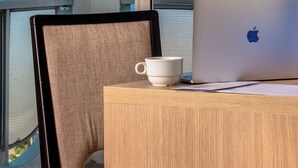 Un ordenador portátil sobre una mesa de madera con una taza blanca, una silla y vistas al océano a través de amplios ventanales.