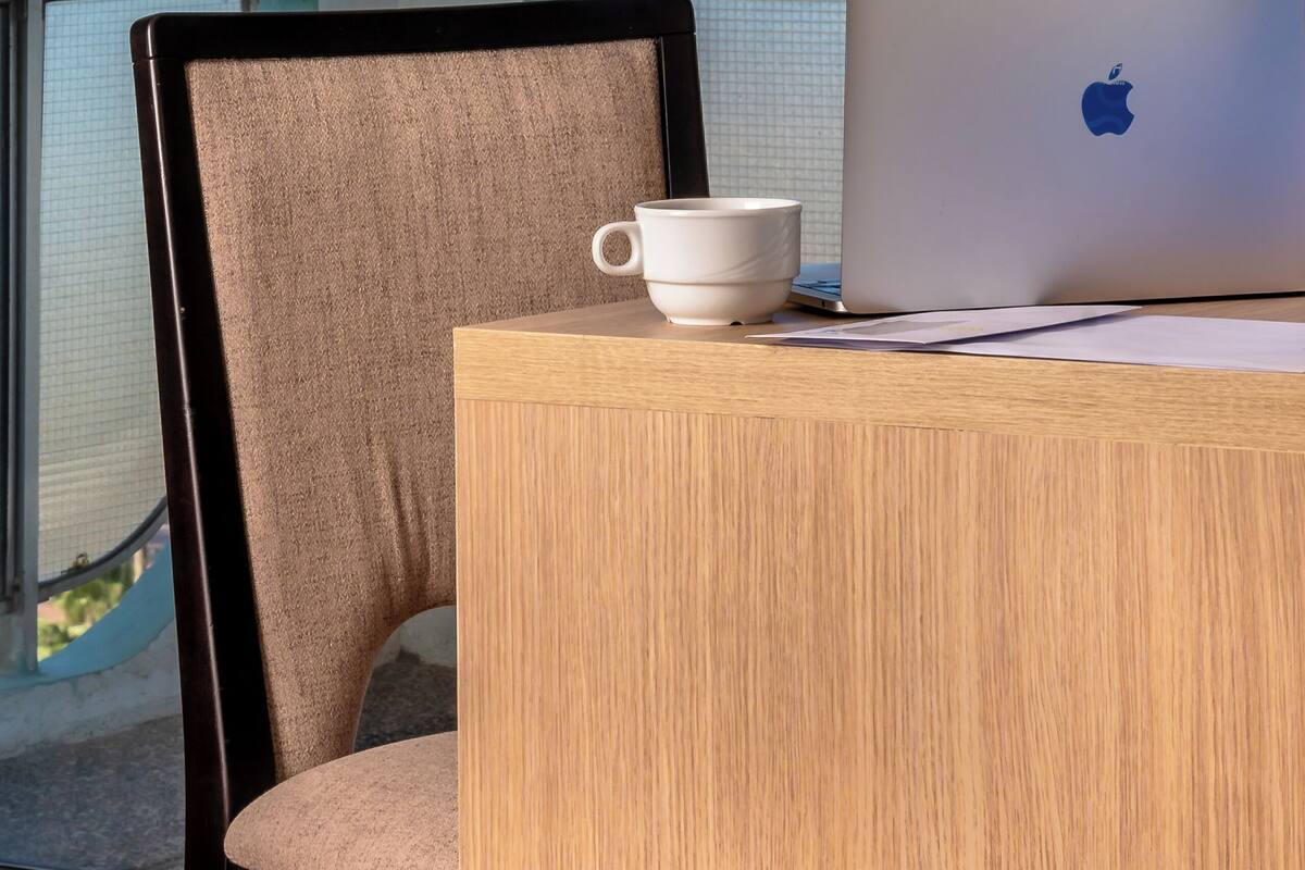 A laptop on a wooden table with a white mug, a chair, and a view of the ocean through large windows.