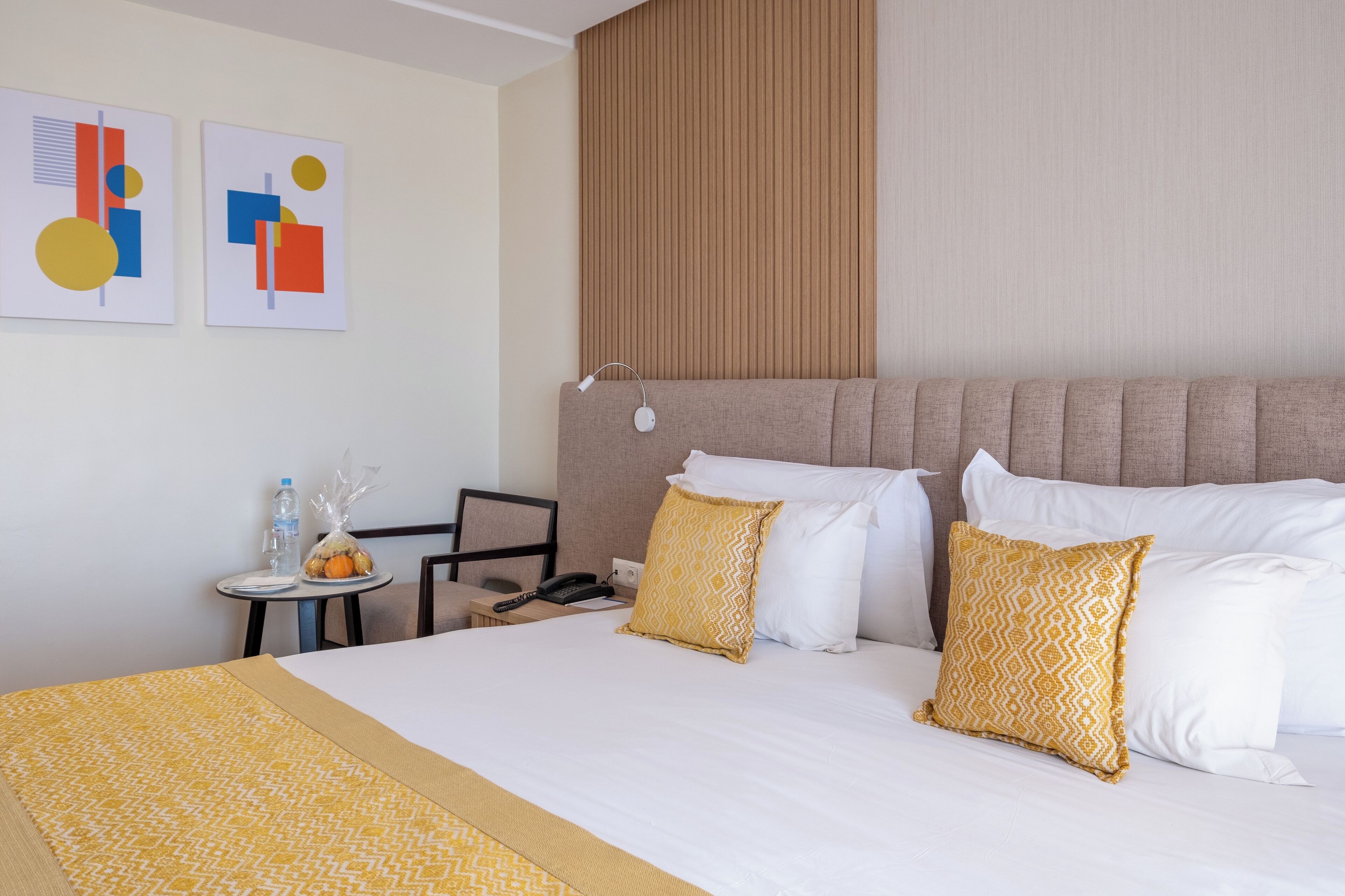 Habitación de hotel con una cama grande, cabecero, una mesita con una silla y dos obras de arte colgadas en la pared.
