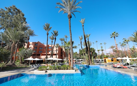 A swimming pool surrounded by palm trees and lounge chairs, with a hotel building in the background.