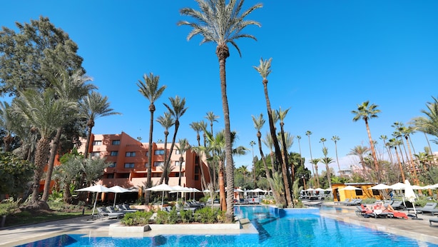 Una piscina rodeada de palmeras y sillas de descanso, con un edificio de hotel al fondo.