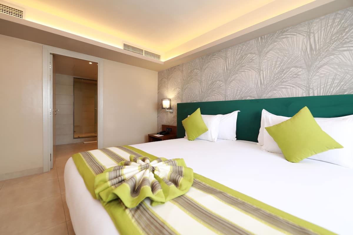 A hotel room with a large bed, a nightstand, a wall-mounted lamp, and a door leading to a bathroom.