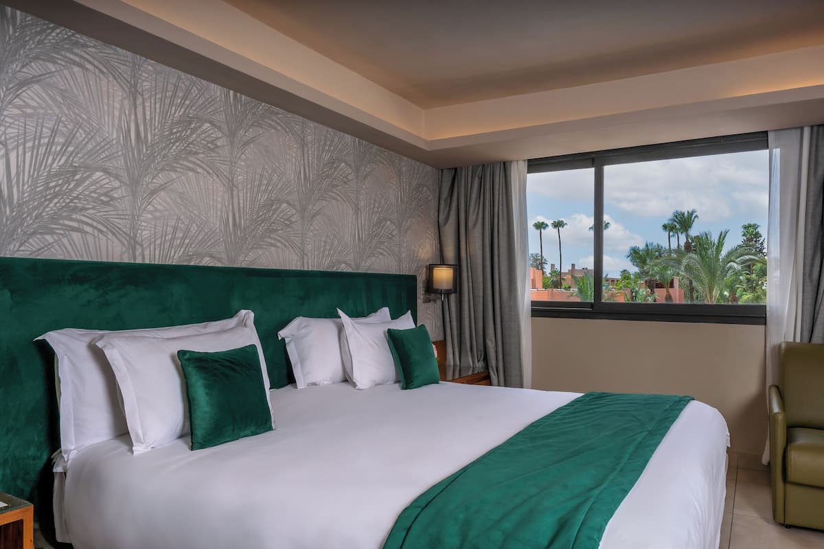 A hotel room with a large bed, a green headboard, white bedding, and a view of palm trees through the window.