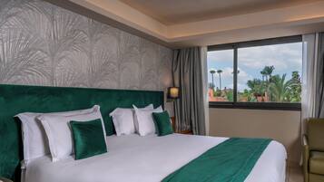 Habitación de hotel con una cama grande, cabecera verde, ropa de cama blanca y vistas a palmeras a través de la ventana.