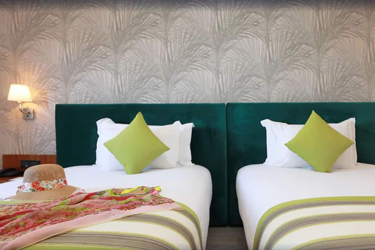 A hotel room with two beds, each with a green headboard and white bedding. There are two bedside lamps and a hat on the left bed.