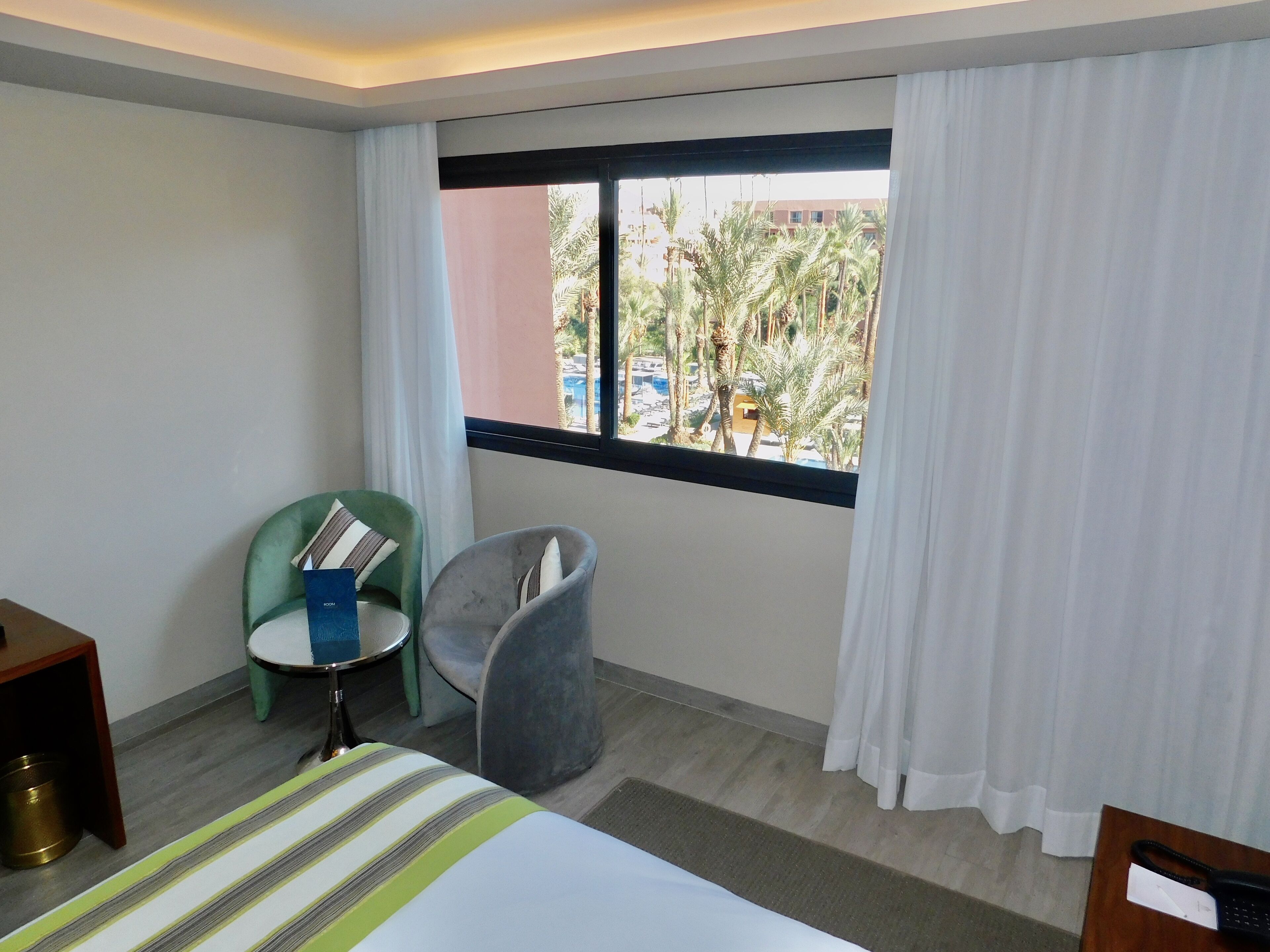 Habitación de hotel con cama, una mesita, dos sillas, ventana con cortinas y vistas a palmeras y edificios.
