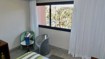 Habitación de hotel con cama, una mesita, dos sillas, ventana con cortinas y vistas a palmeras y edificios.