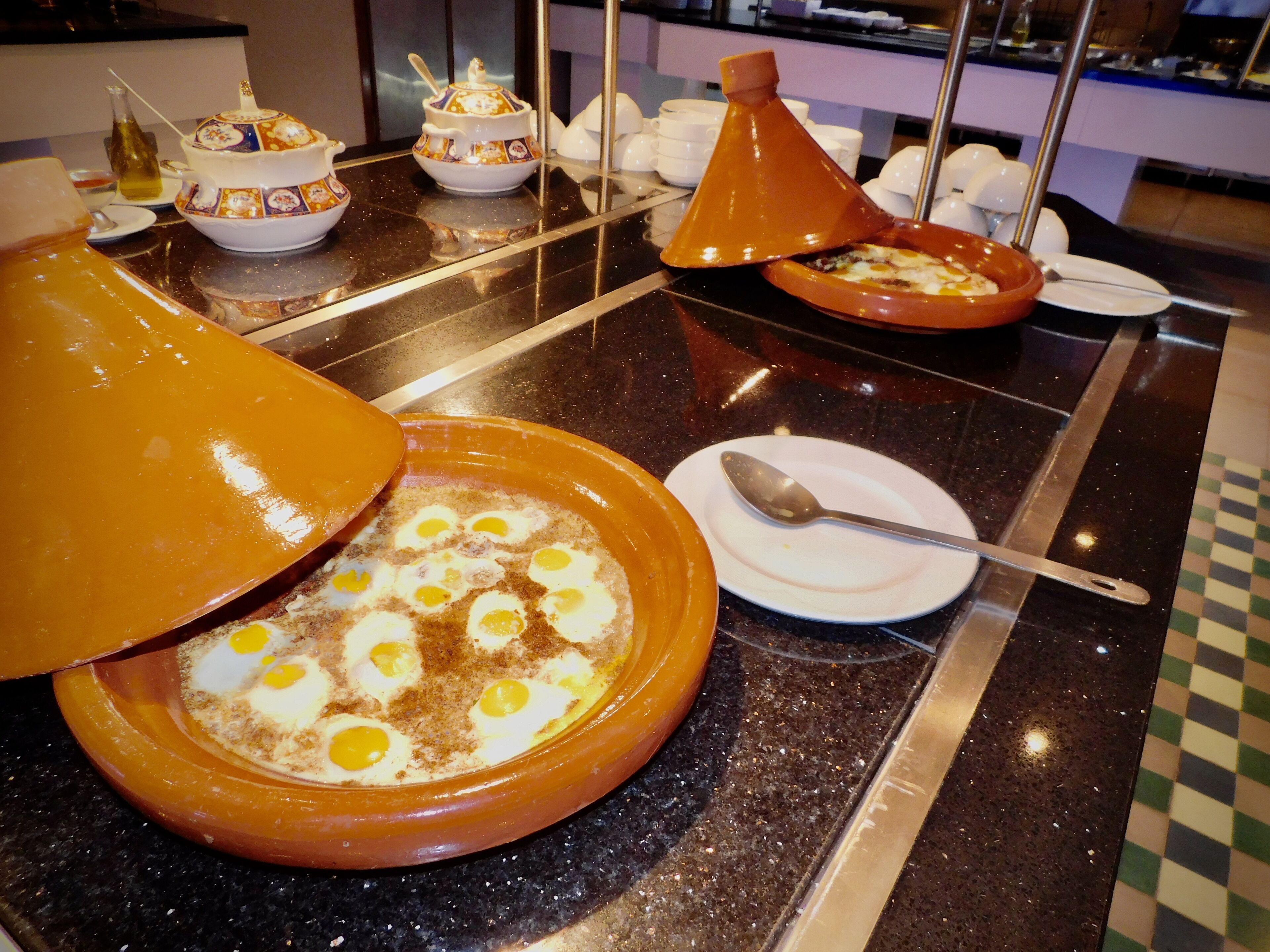 Un bufé de desayuno con diversos platos, incluyendo un tajín con huevos, una botella de aceite de oliva y varias teteras.