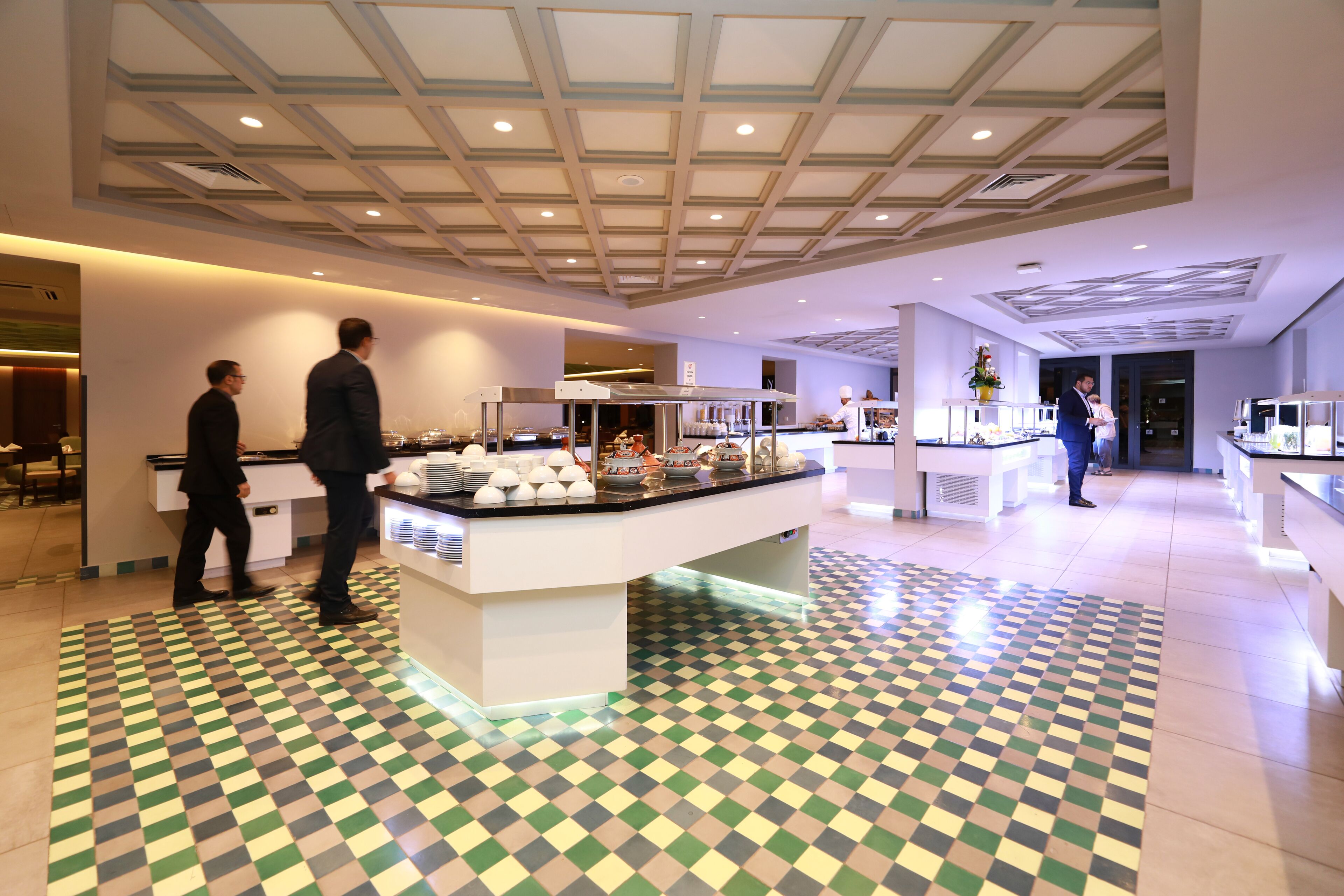 A modern hotel lobby with a checkered floor, a central island with food displays, and a ceiling with recessed lighting.
