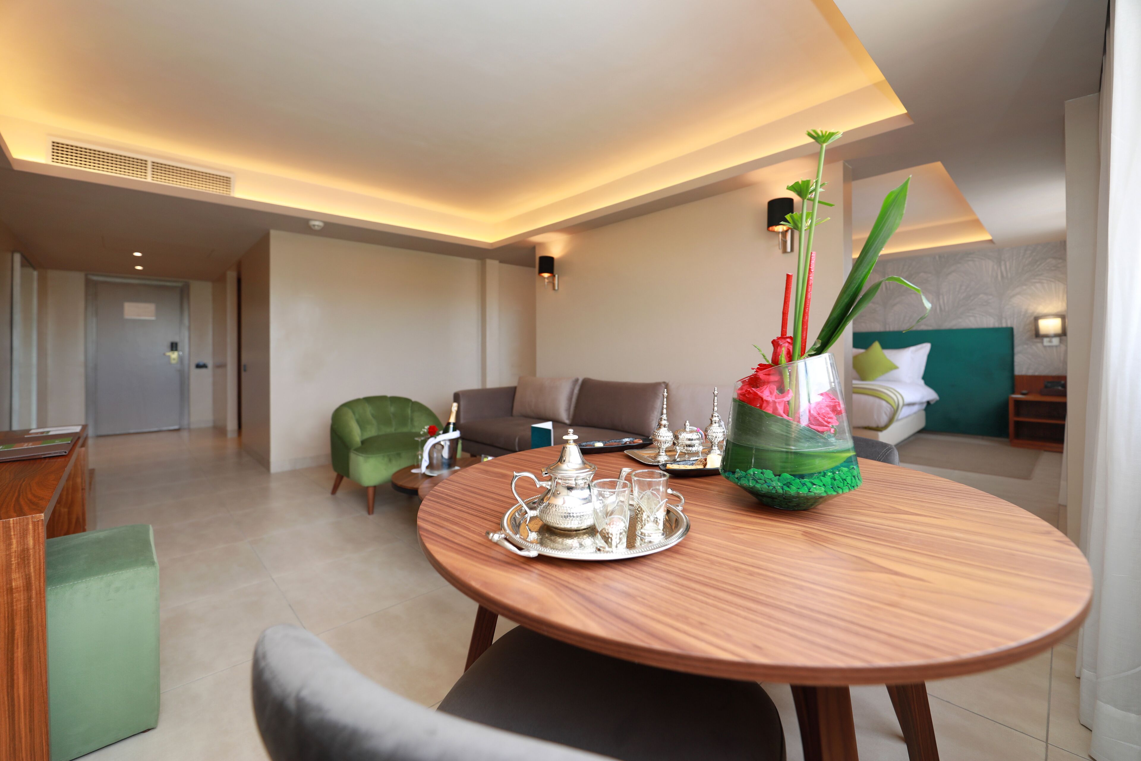 Ein modernes Hotelzimmer mit Essbereich, der einen Holztisch mit Besteck und Tablett, einen grünen Sessel und eine Couch umfasst.