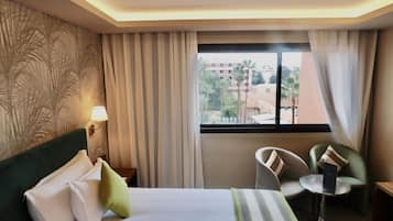 Une chambre d’hôtel comprenant un lit, une table de chevet, une lampe, une chaise, une petite table et une fenêtre donnant sur des immeubles et des palmiers.