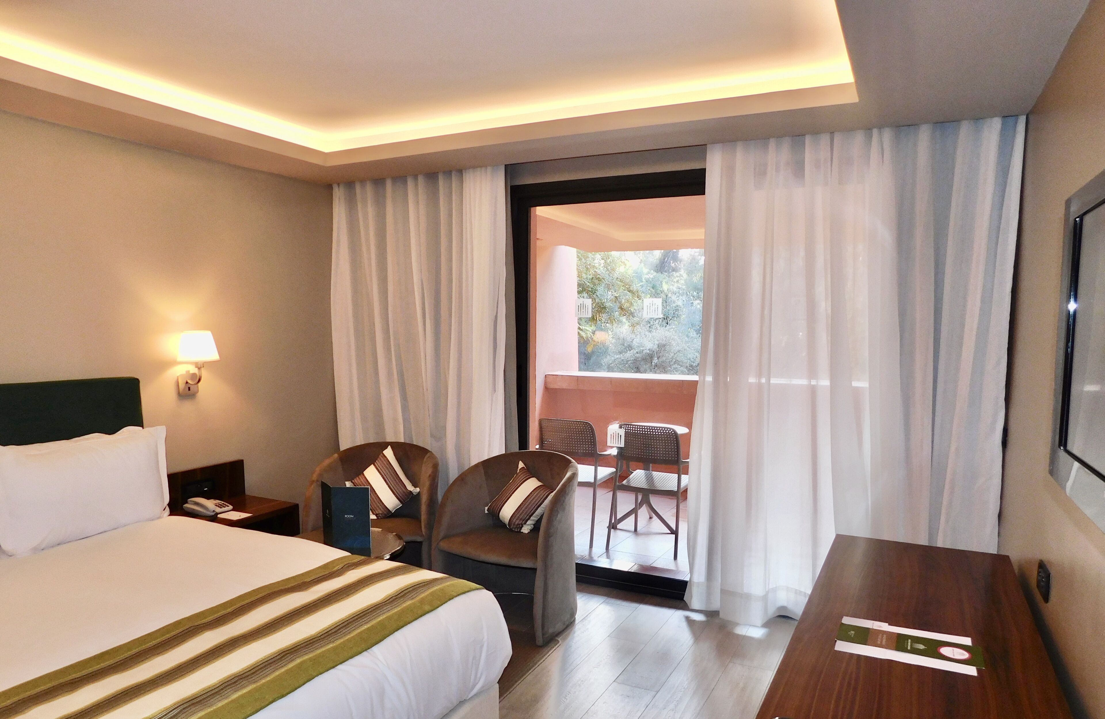 A hotel room with a large bed, a desk, two chairs, a balcony with a view, and a wall-mounted TV.