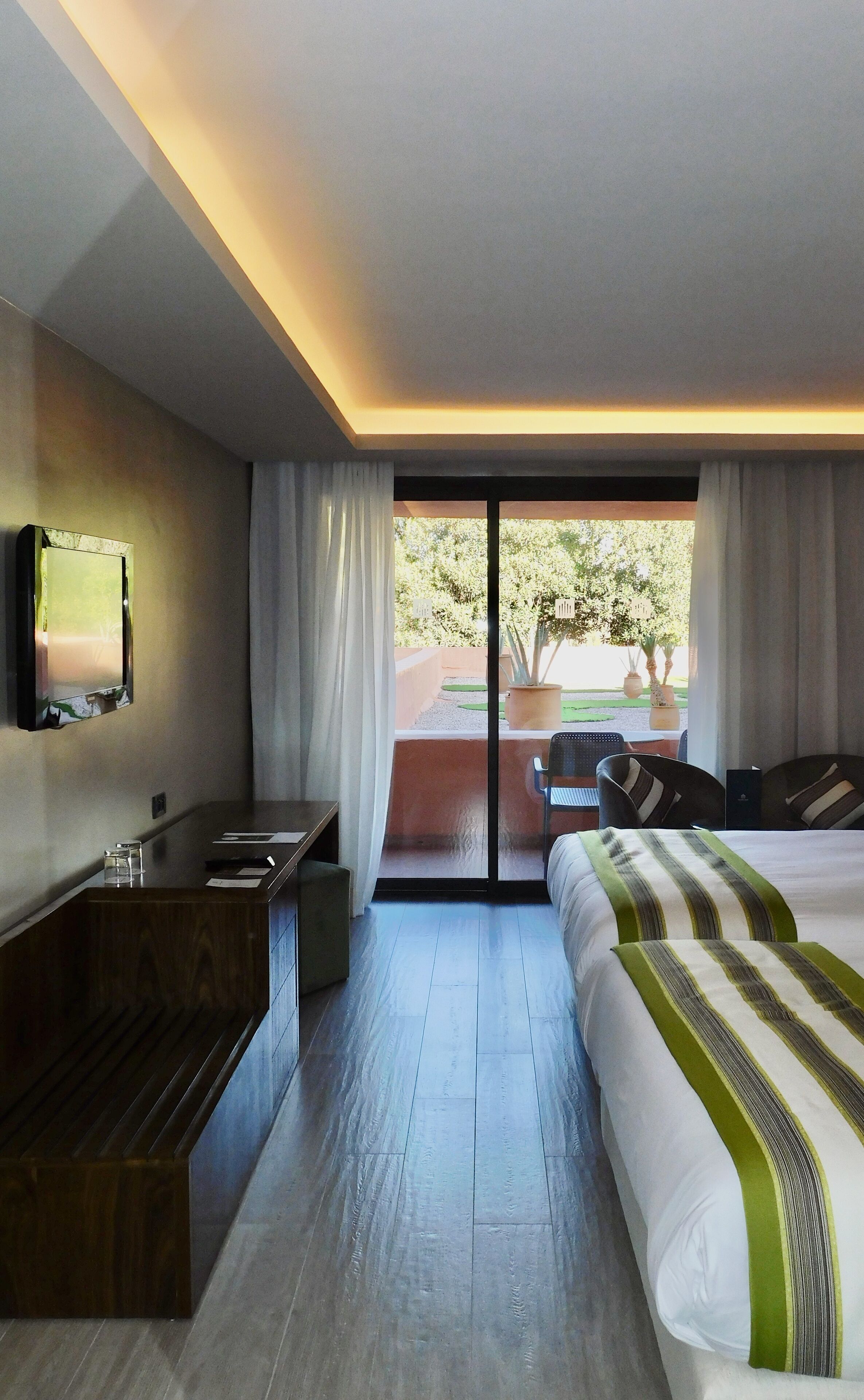 A hotel room with a bed, a desk, a TV, and a balcony with a view of trees.
