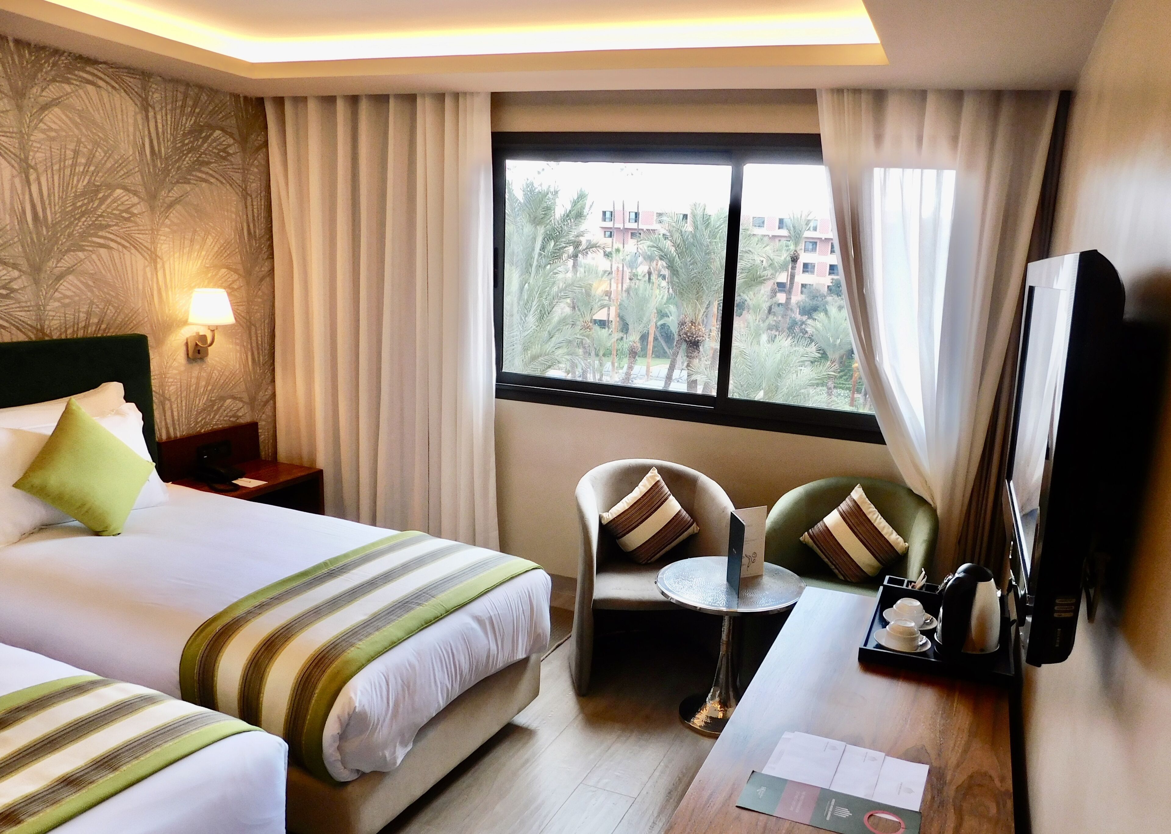 A hotel room with two beds, a small table, a chair, a TV, and a window with a view of palm trees and buildings.