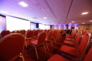 Una sala conferenze con file di sedie rosse, un grande schermo e un palco con un leggio.