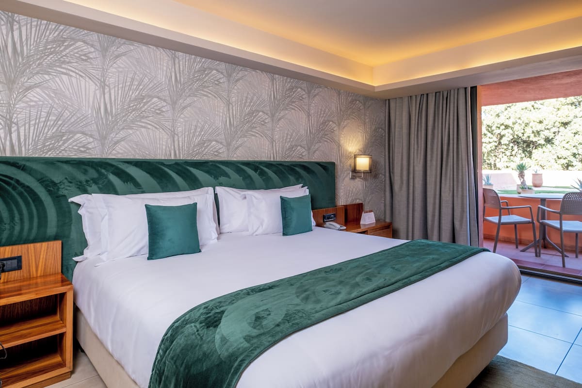 A large bed with a green headboard, white bedding, and a green throw blanket. There is a wooden nightstand with a phone and a lamp on it. The room has a large window with curtains and a view of the outside.