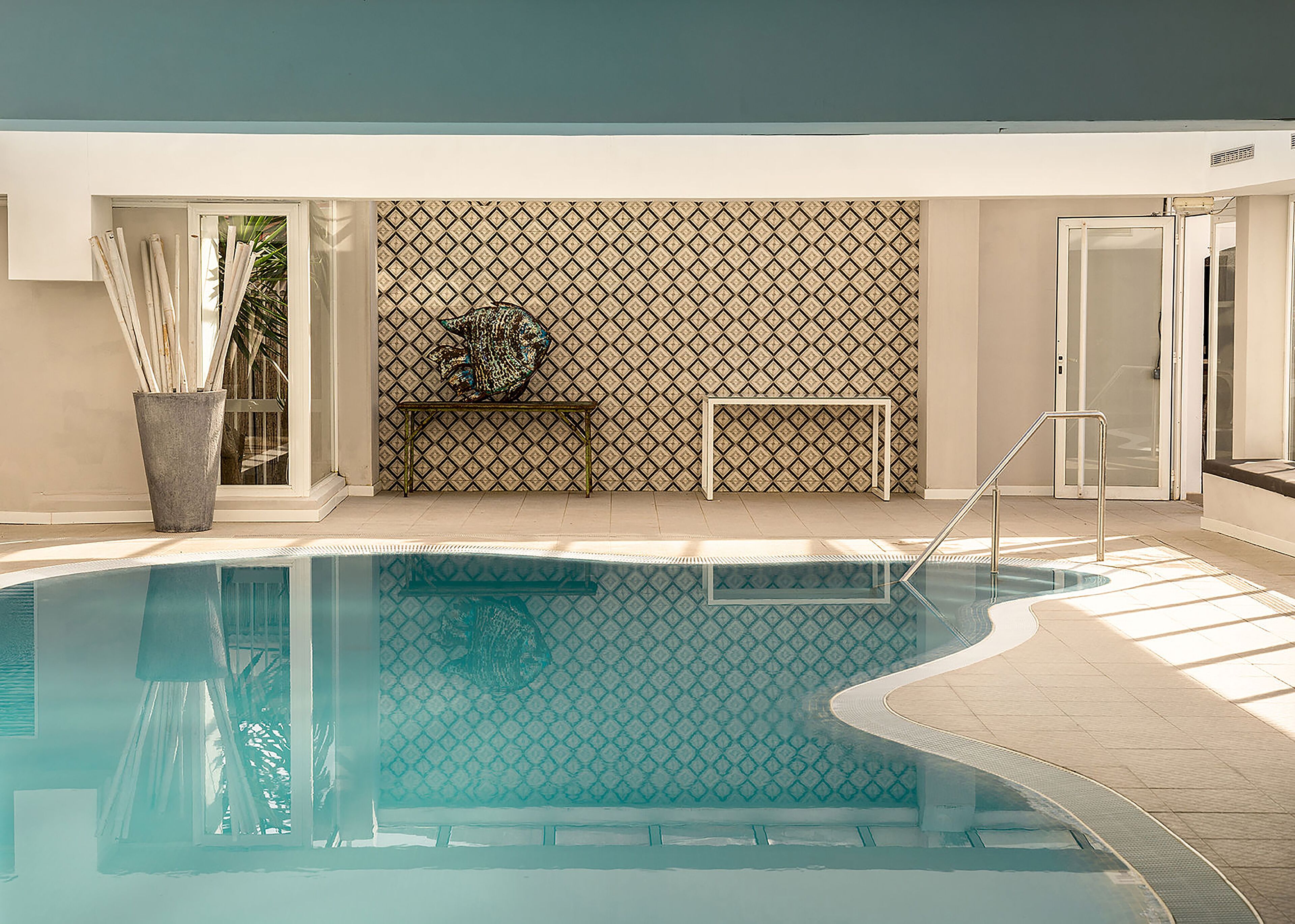Une piscine avec un mur orné d’un motif géométrique, une petite table et une plante en pot.