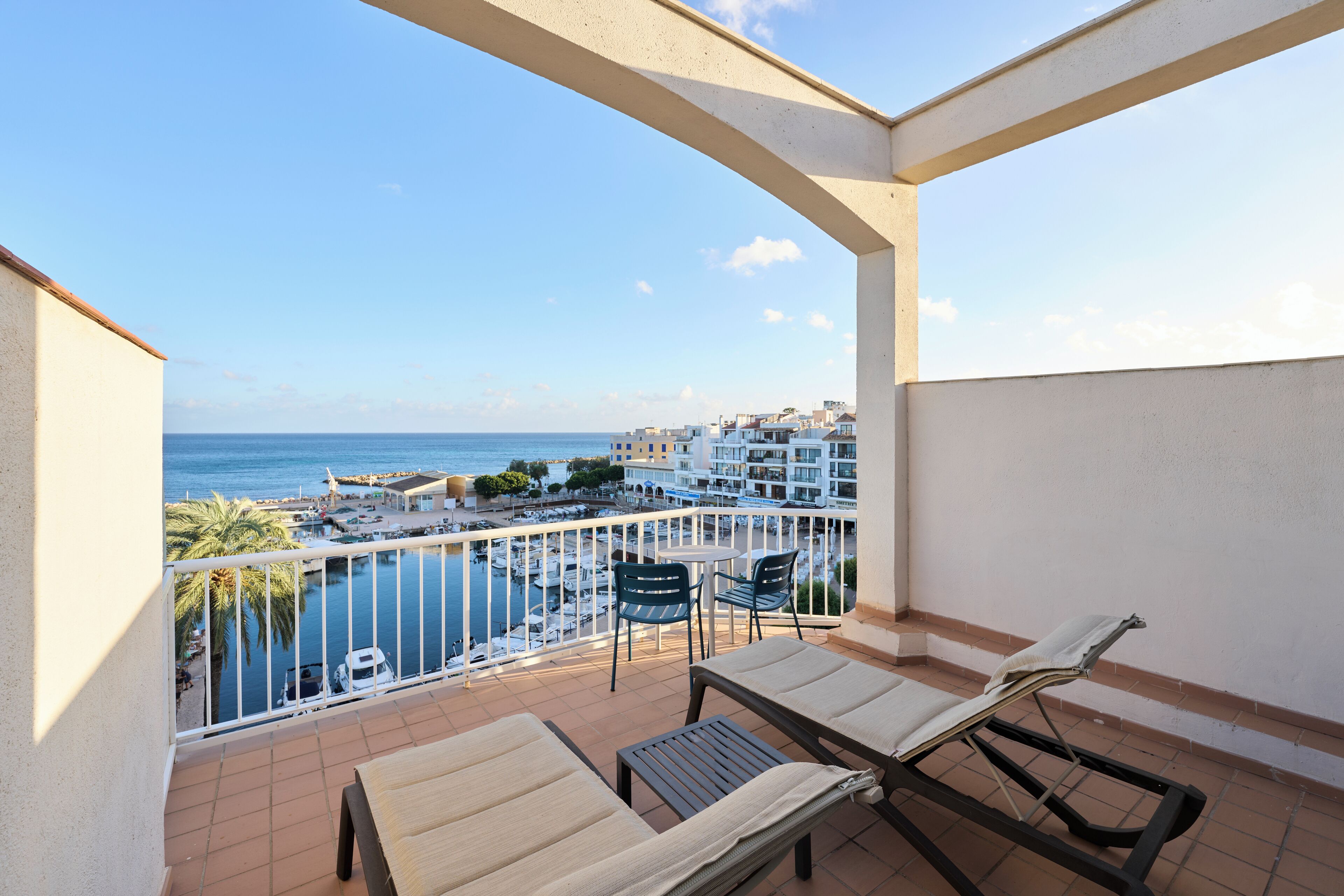 A balcony with two lounge chairs and a table, overlooking a marina and buildings.