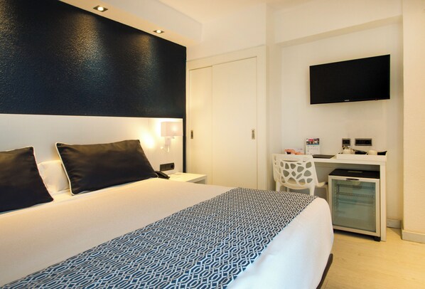 A hotel room with a large bed, a desk with a computer, a television, and a minibar. - Catalonia Del Mar - Adults Only (Son Servera)