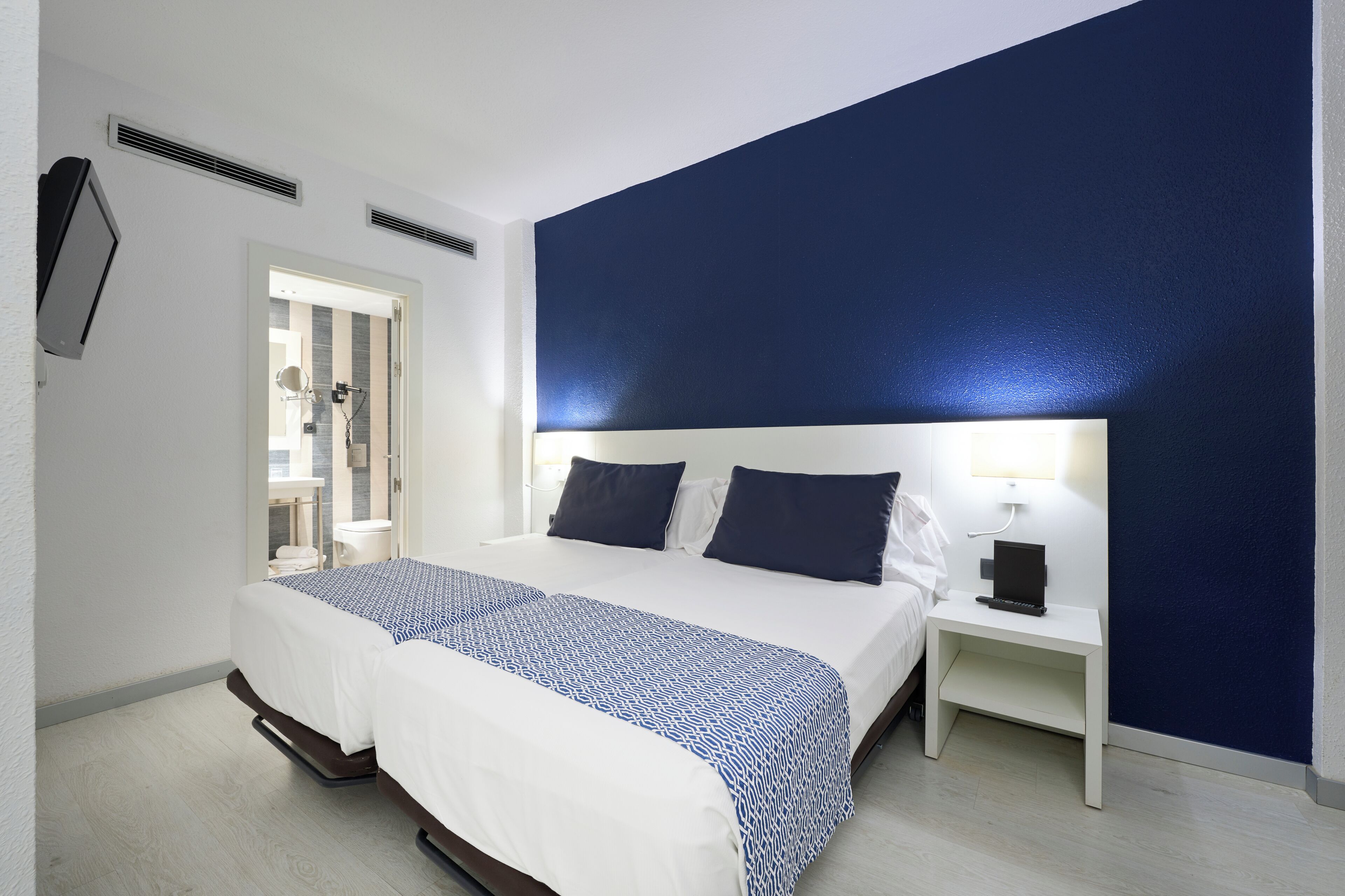 Habitación de hotel con una cama grande, una mesita de noche con lámpara y vista al baño a través de una puerta abierta.