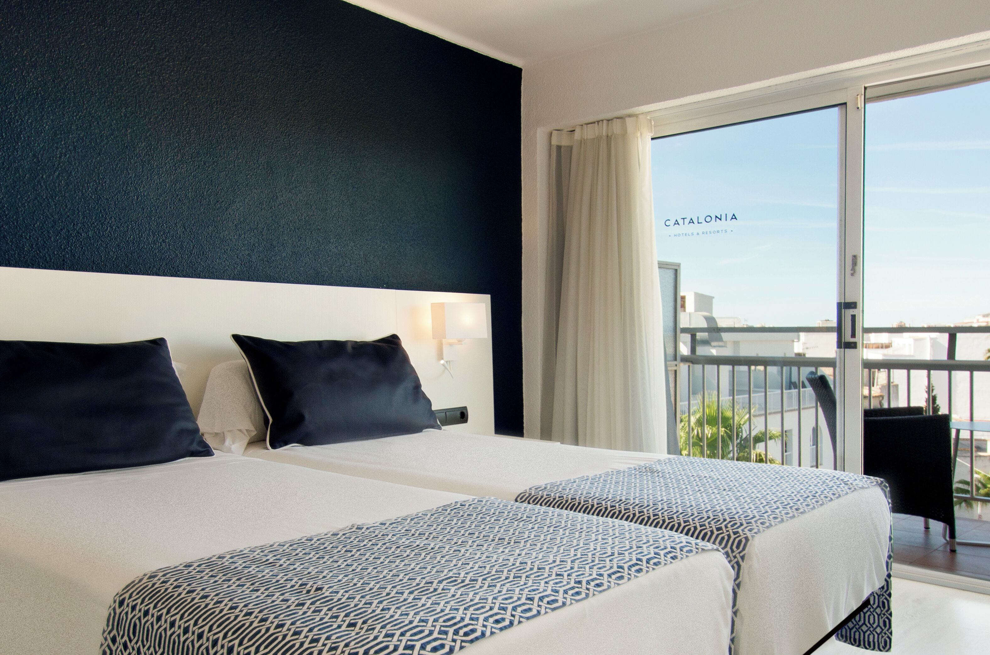 A hotel room with two beds, a balcony with a view of the city, and a sign reading "Catalonia".