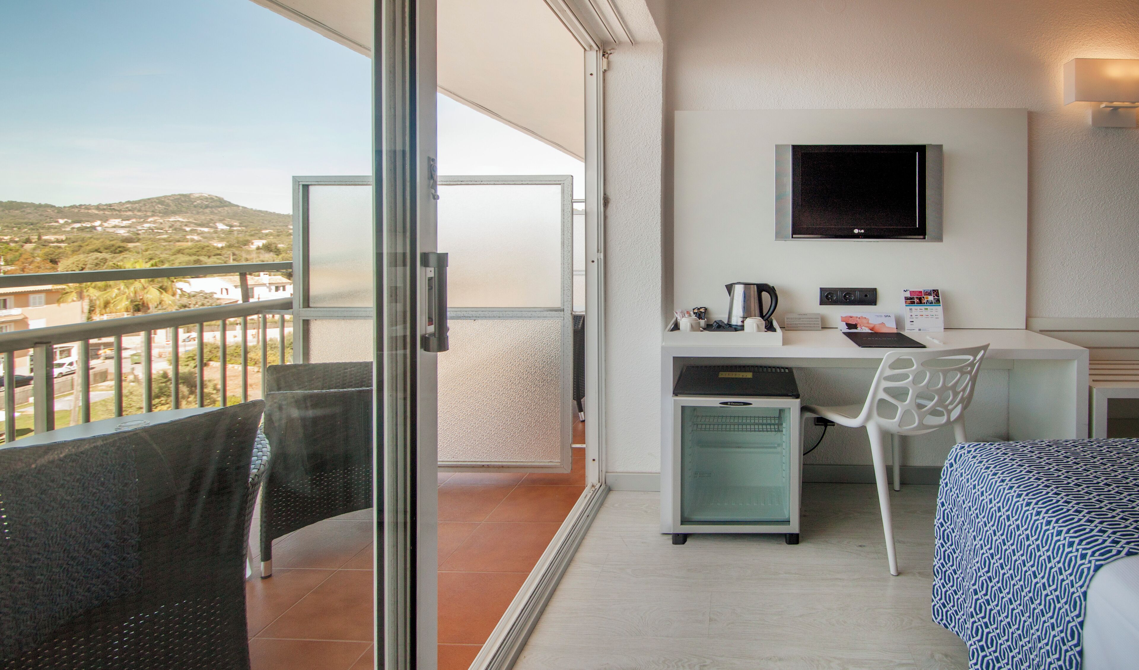 A modern hotel room with a balcony, a desk with a chair, a television, and a minibar.