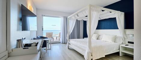 A modern hotel room with a four-poster bed, a desk with a chair, a flat-screen TV, and a balcony with a view.