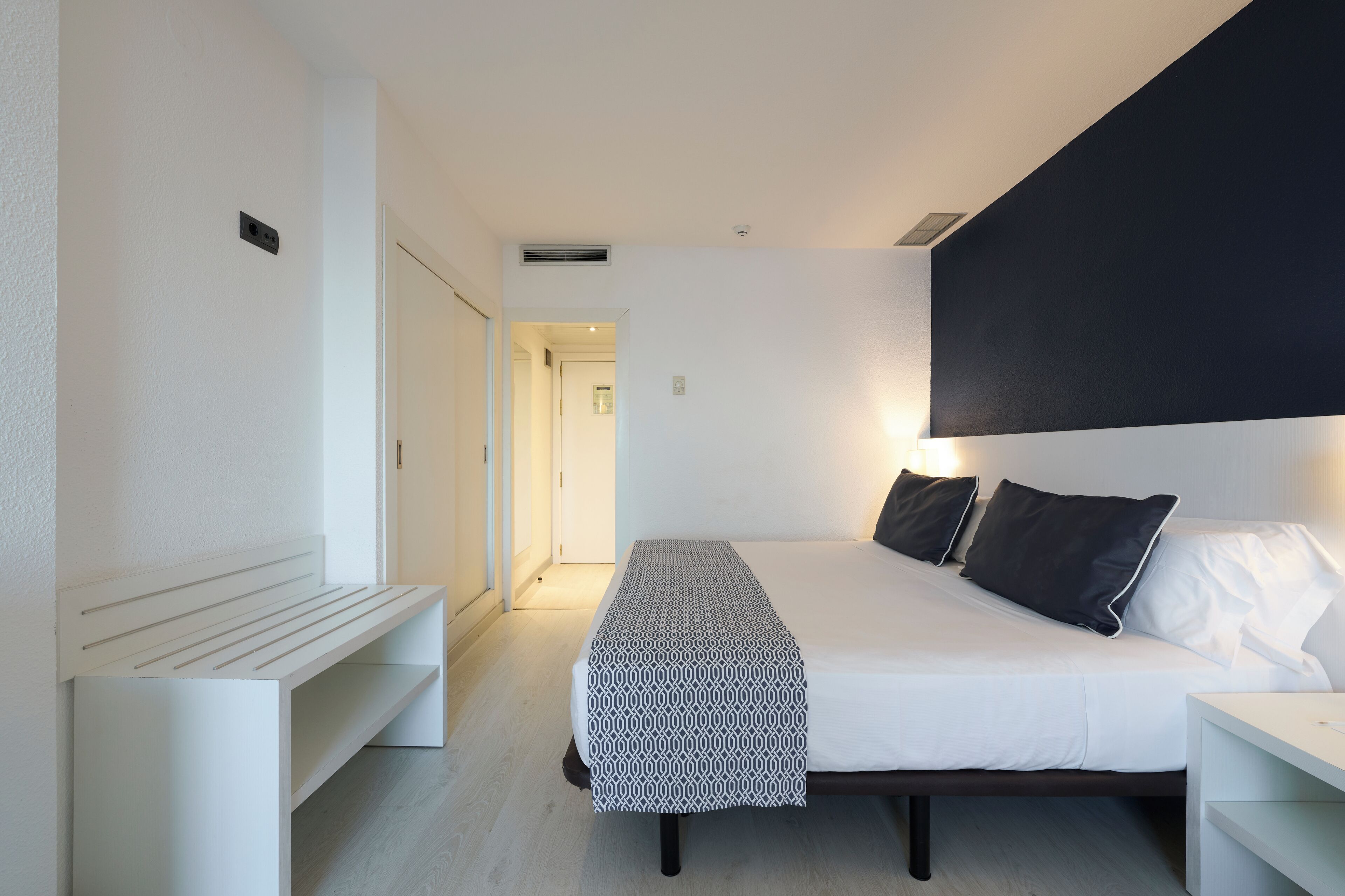 A modern hotel room with a large bed, bedside tables, a bench, and a dark accent wall.