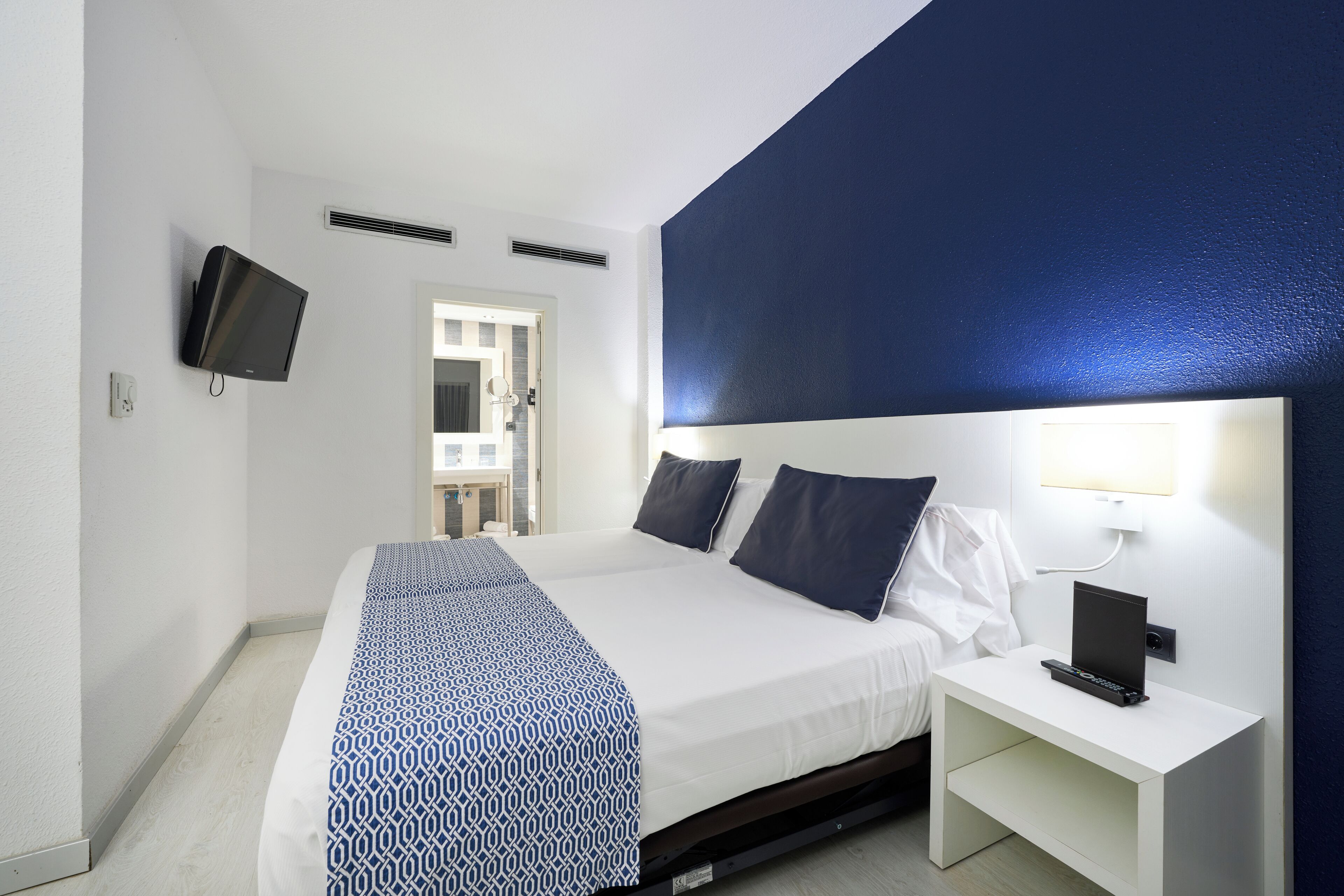 A hotel room with a bed, a TV mounted on the wall, a bedside table with a lamp, and a door leading to a bathroom.