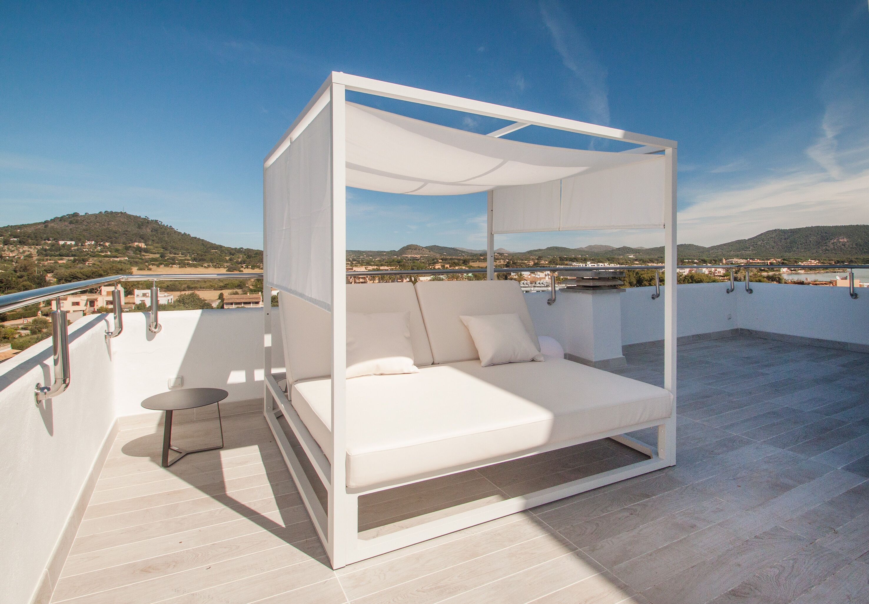 A white four-poster bed with a table on a balcony with a view of a hilly landscape.