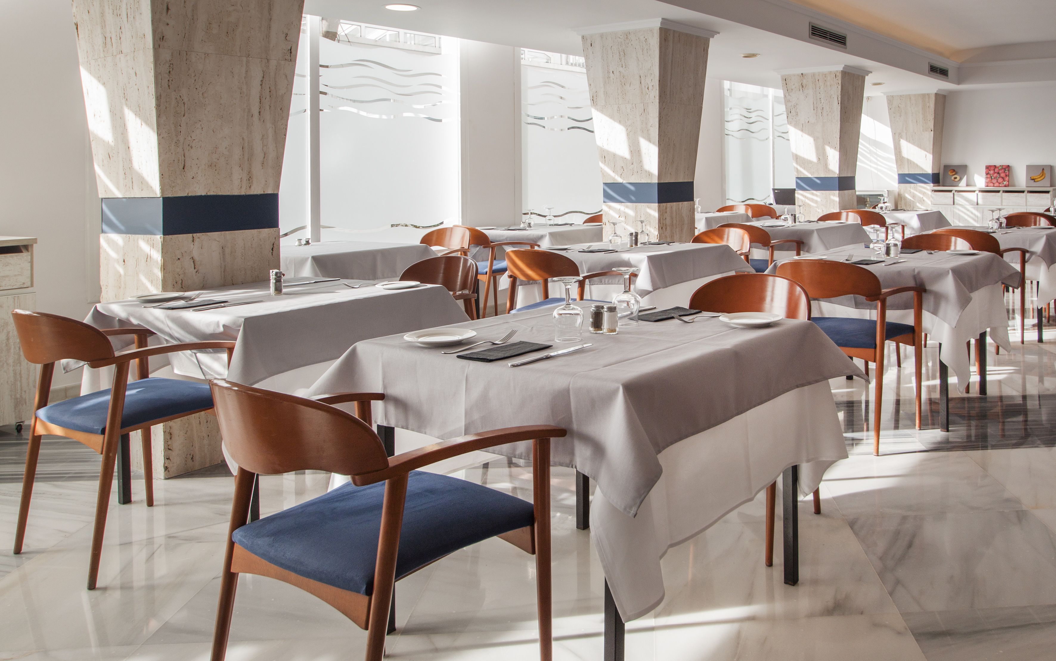 A restaurant with marble floors and white tablecloths, wooden chairs with blue cushions, and large windows allowing natural light.