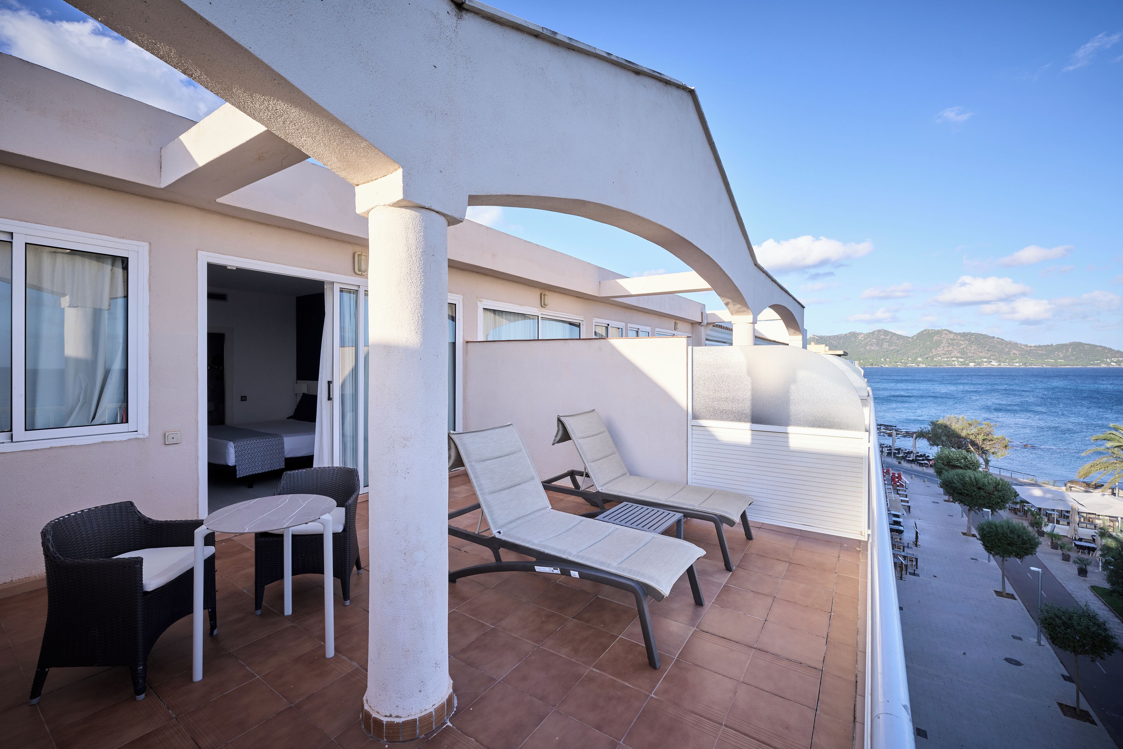 A balcony with two lounge chairs and a small table, overlooking a beach and the sea.