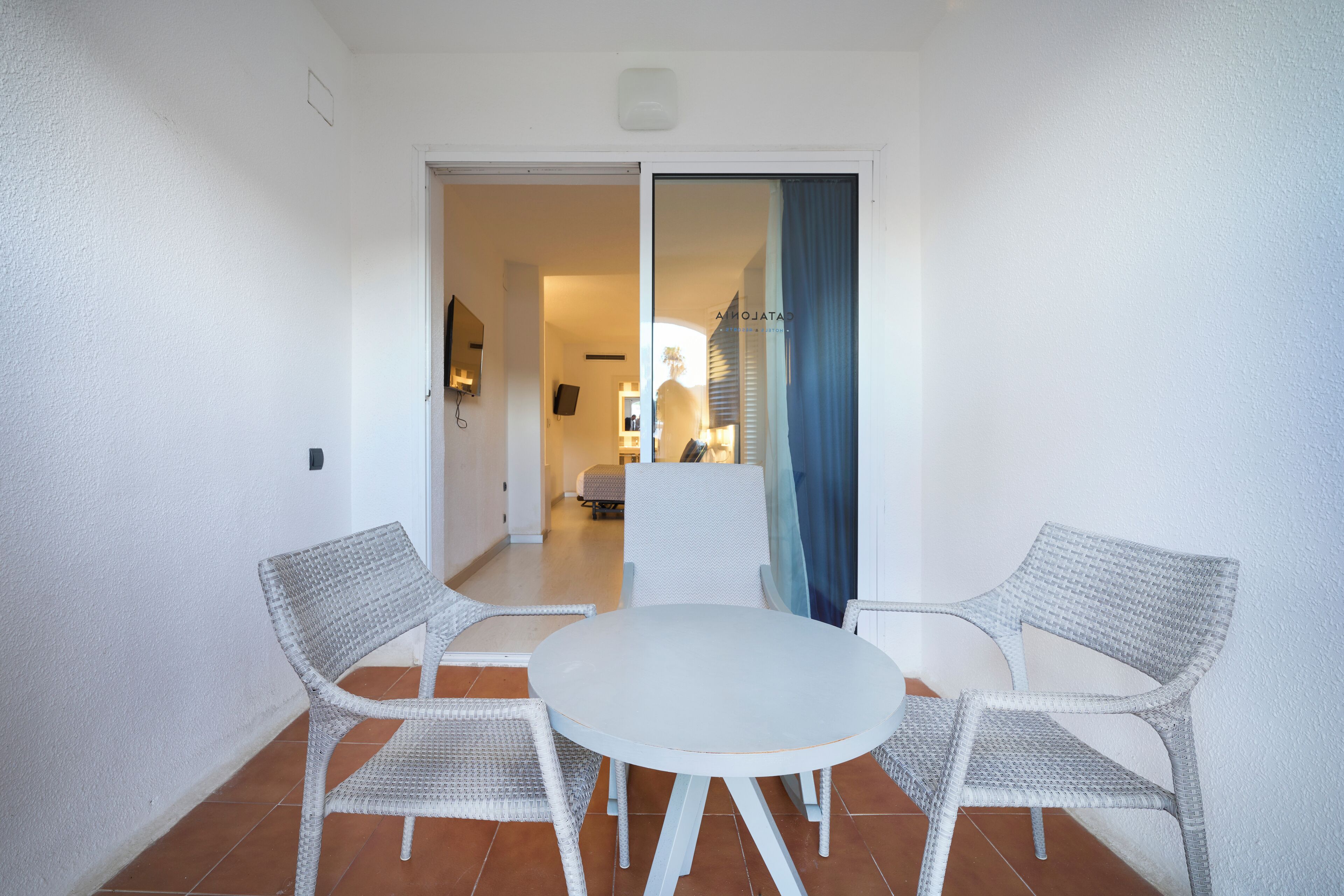 A room with a round table and two wicker chairs, a white wall, and a view into another room with a bed and a window.