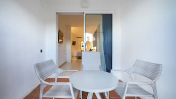 A room with a round table and two wicker chairs, a white wall, and a view into another room with a bed and a window.