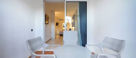 A room with a round table and two wicker chairs, a white wall, and a view into another room with a bed and a window.
