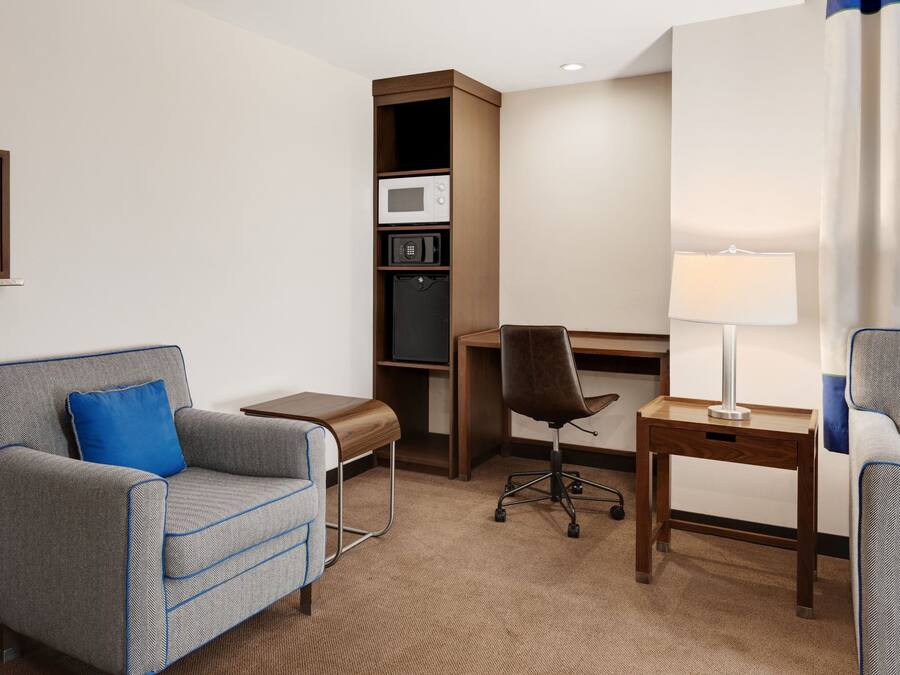 Una habitación de hotel con un sillón gris con detalles azules, un escritorio de madera, una computadora, un refrigerador pequeño y una lámpara.