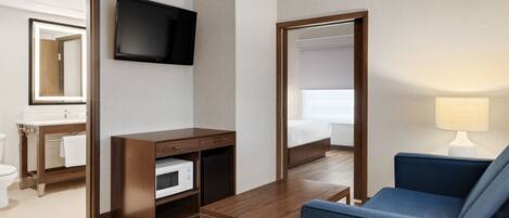 Habitación de hotel con escritorio de madera, sofá azul, televisor montado en la pared, espejo y mesita de noche con lámpara.