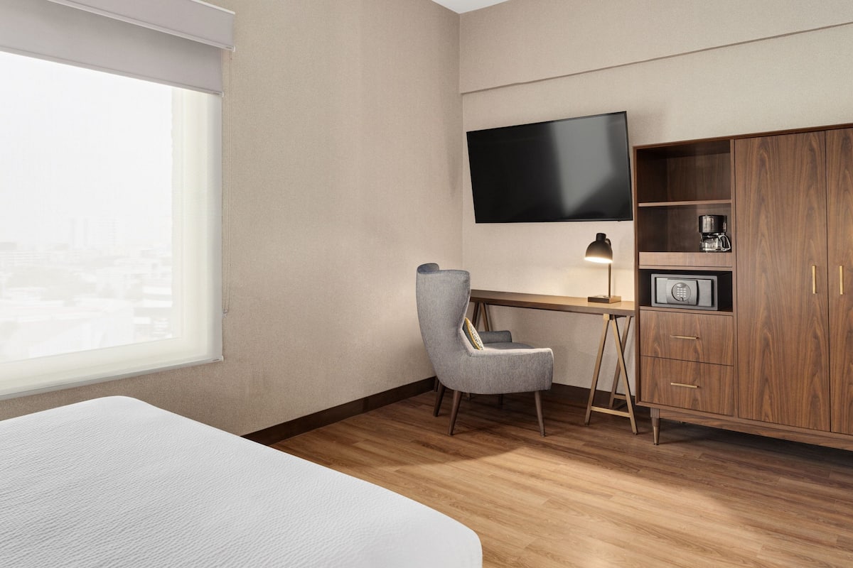 Une chambre d’hôtel comprenant un lit, un bureau avec une chaise, un téléviseur fixé au mur, une armoire en bois et une machine à café.