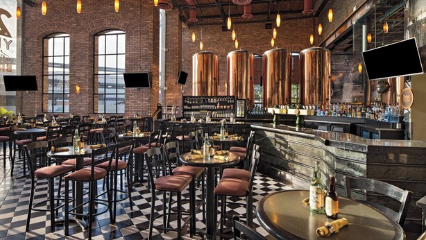 A spacious restaurant with high ceilings, exposed brick walls, and a checkered floor. Tables and chairs are arranged for dining, and a bar area is visible with a variety of bottles and glasses.