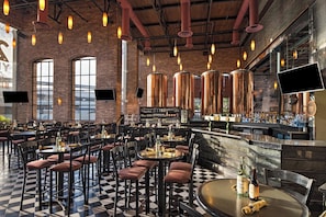 A spacious restaurant with high ceilings, exposed brick walls, and a checkered floor. Tables and chairs are arranged for dining, and a bar area is visible with a variety of bottles and glasses.