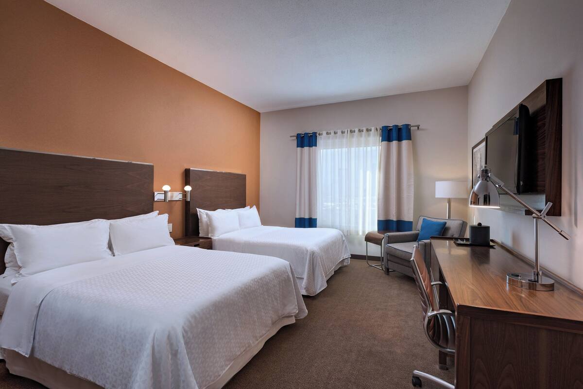 Une chambre d’hôtel avec deux lits, un bureau, une chaise, une télévision et une fenêtre avec des rideaux.