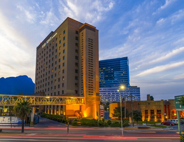 A hotel with a prominent sign, a modern building across the street, and a bridge overhead.