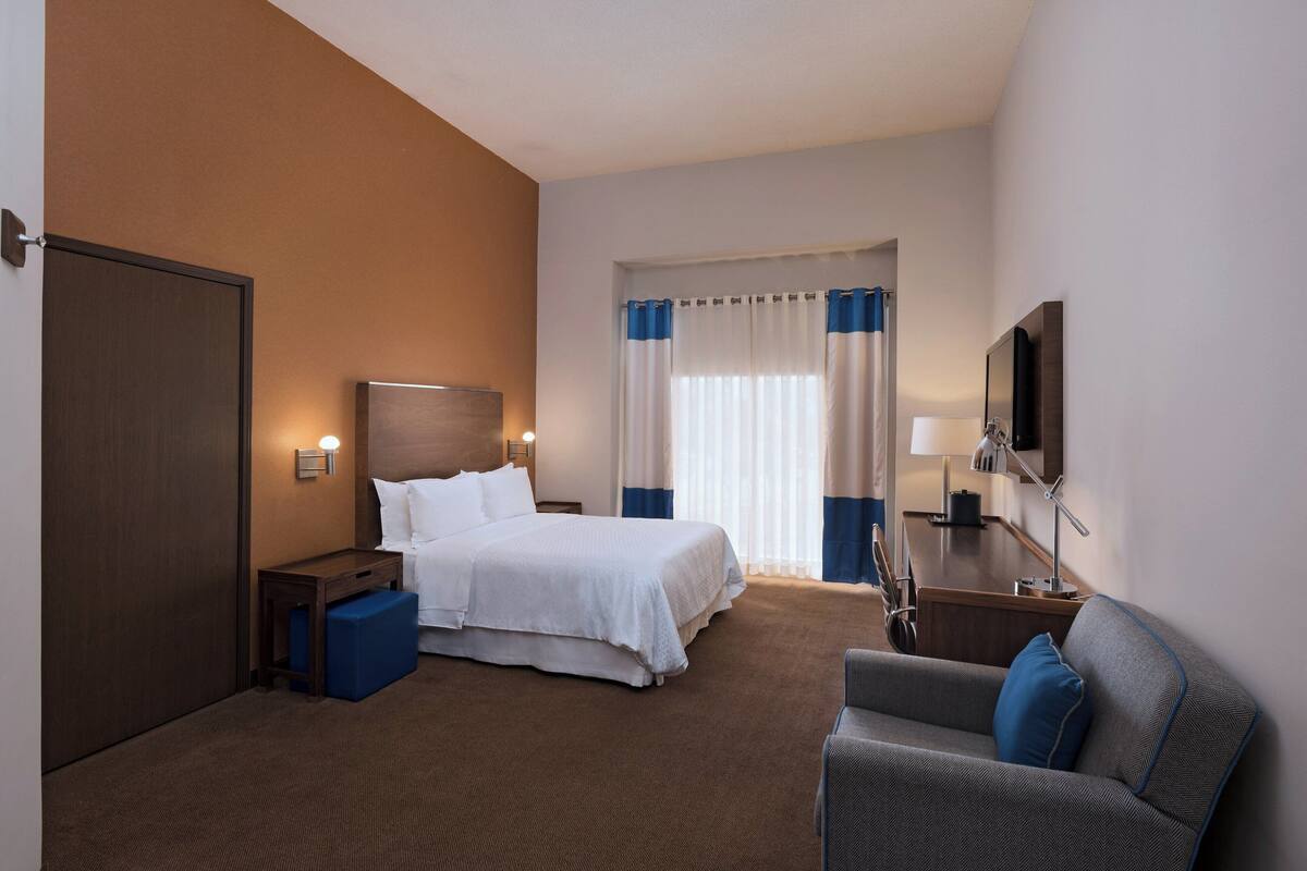 Une chambre d’hôtel avec un lit, un bureau, une chaise, une télévision et une fenêtre avec des rideaux.