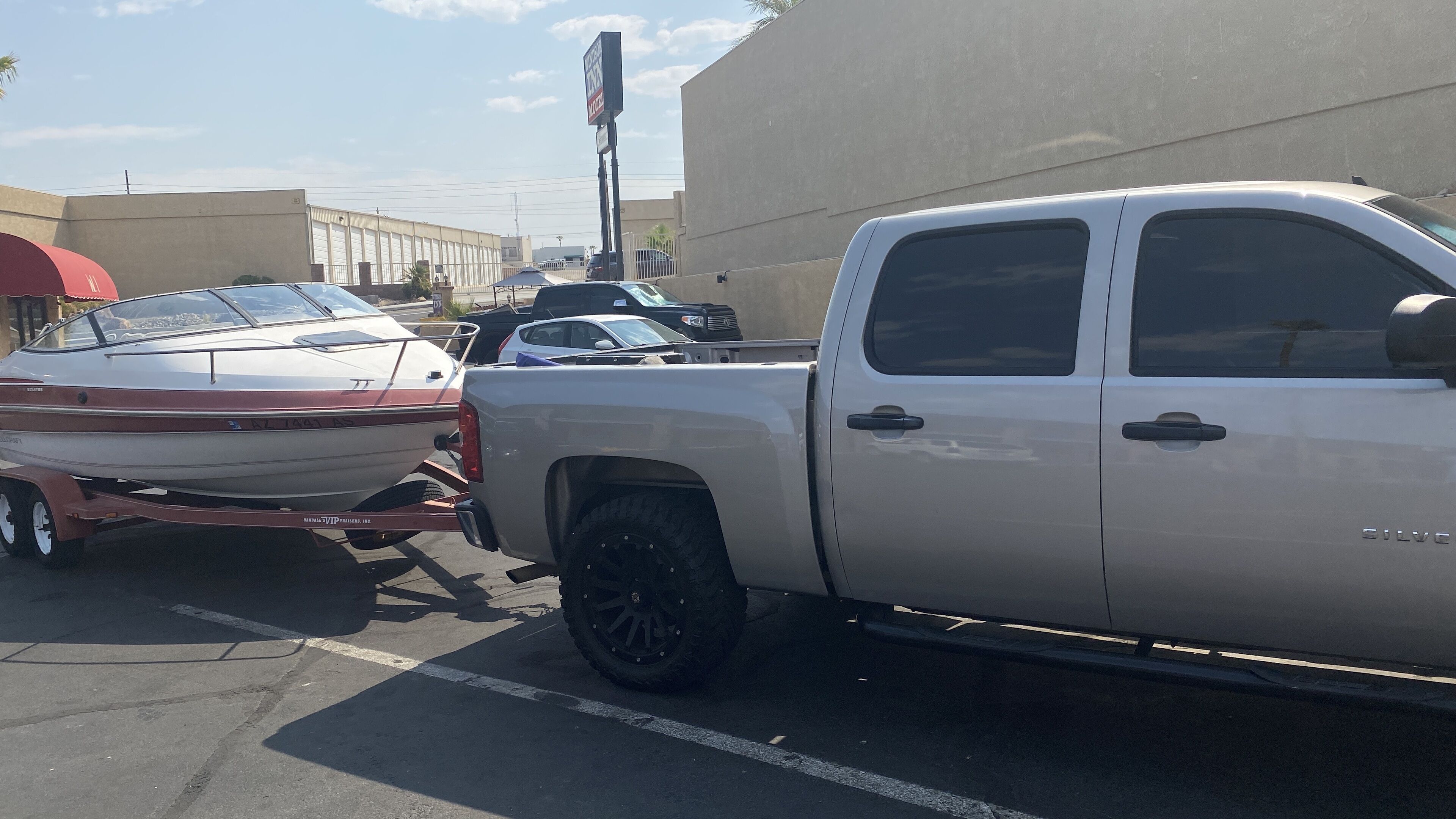 A pickup truck parked in a lot with a trailer attached, a boat on the trailer, and a building in the background.