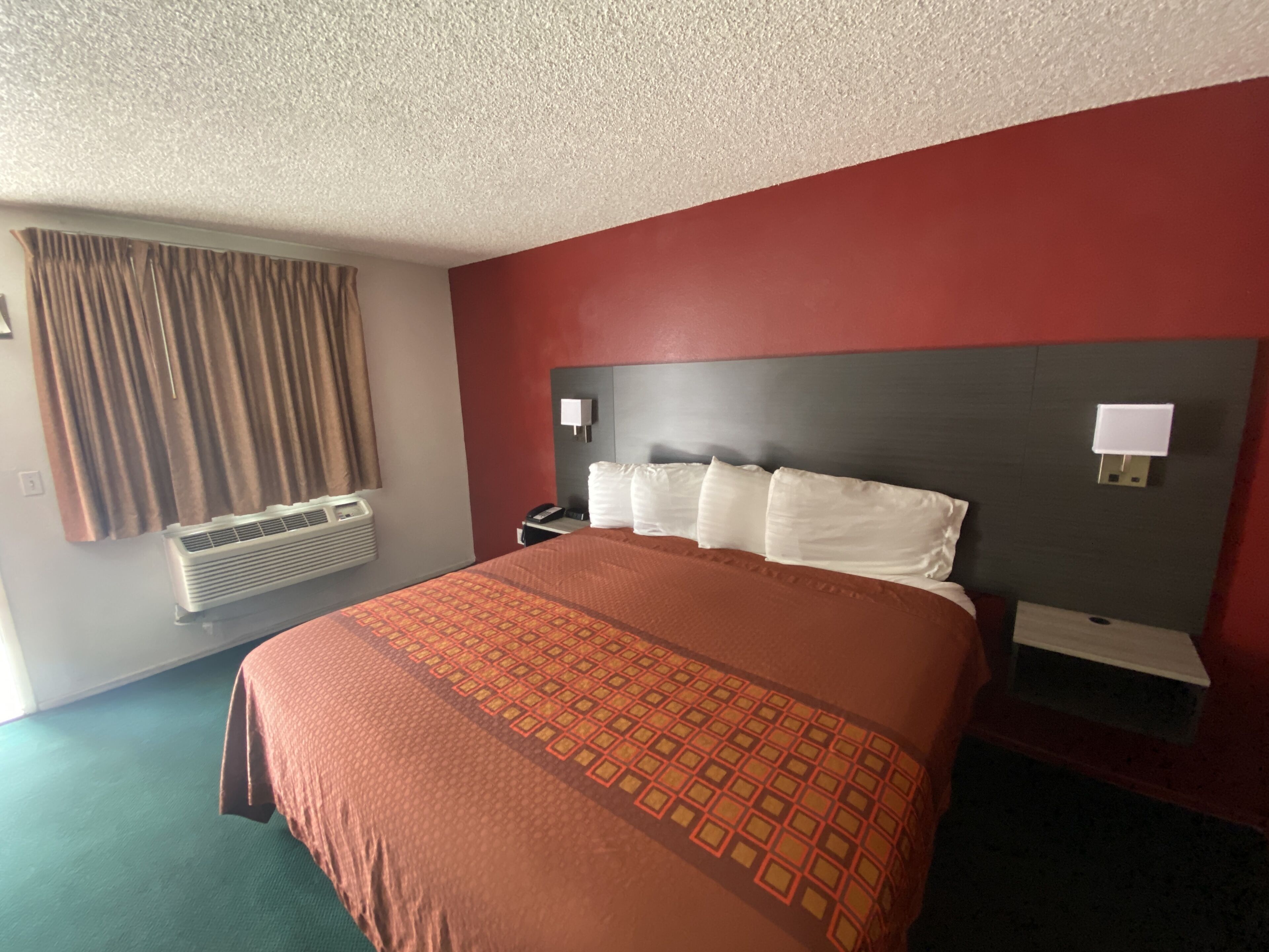 A hotel room with a large bed, a headboard with two wall-mounted lamps, a bedside table, and a window with curtains.