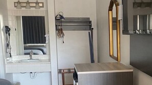 A compact hotel room with a bed, a small desk, a mirror, a sink, and a shower area.