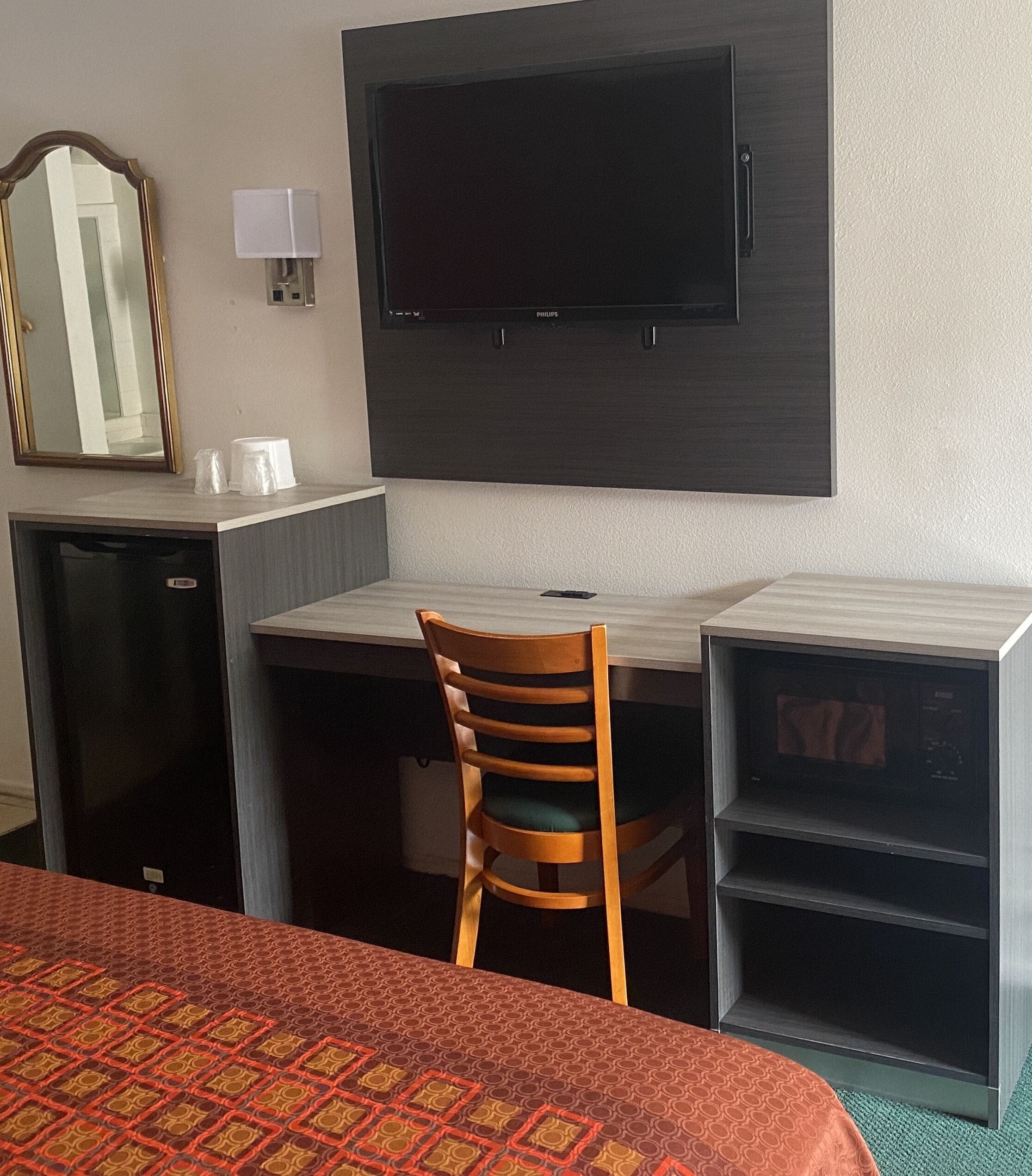 A hotel room with a bed, a desk with a chair, a television, a mirror, and a small refrigerator.