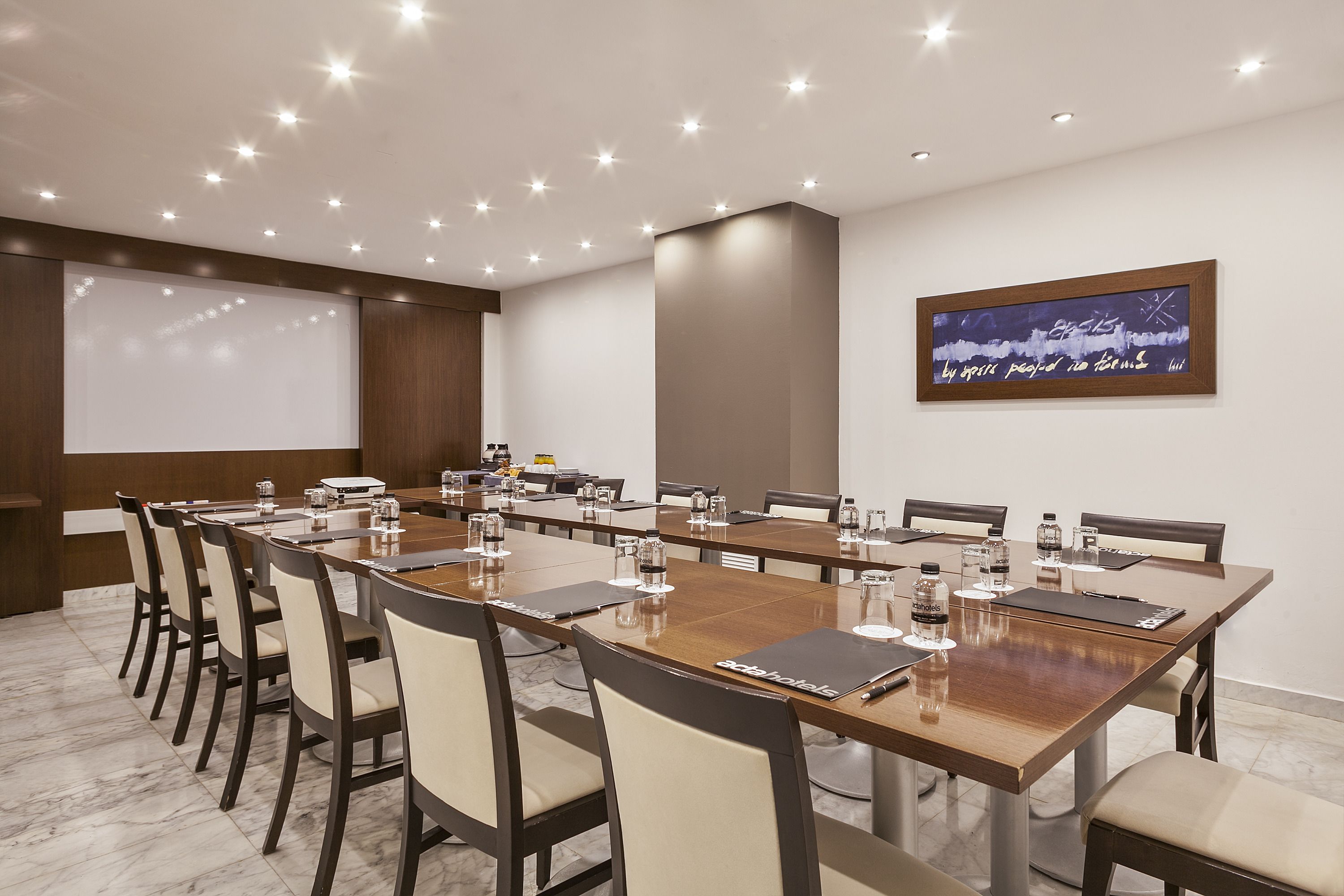 A conference room with a long table, chairs, a projector screen, and a wall-mounted display.