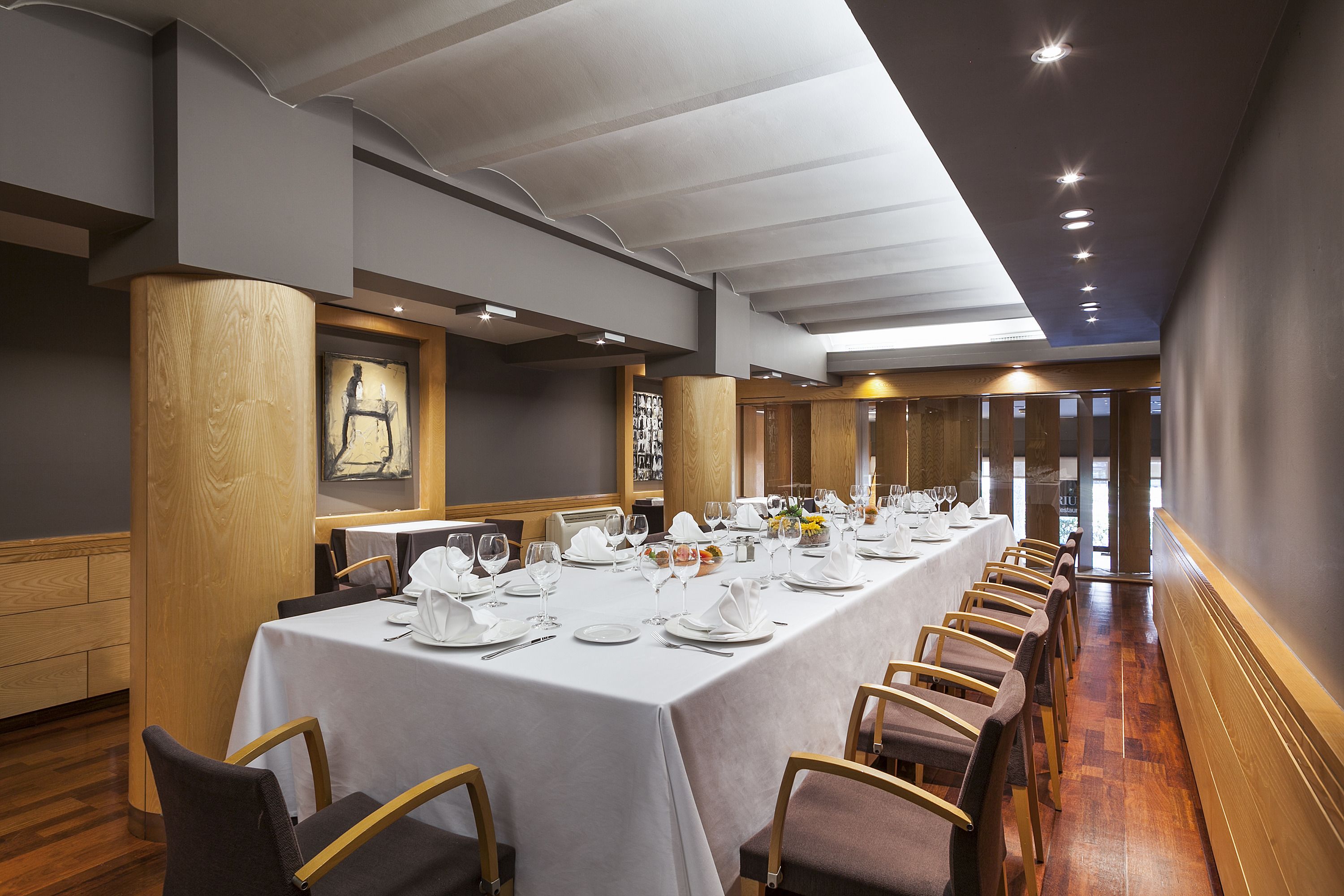 A long dining table set with white tablecloths, chairs, and dinnerware, in a modern interior with wooden accents and recessed lighting.